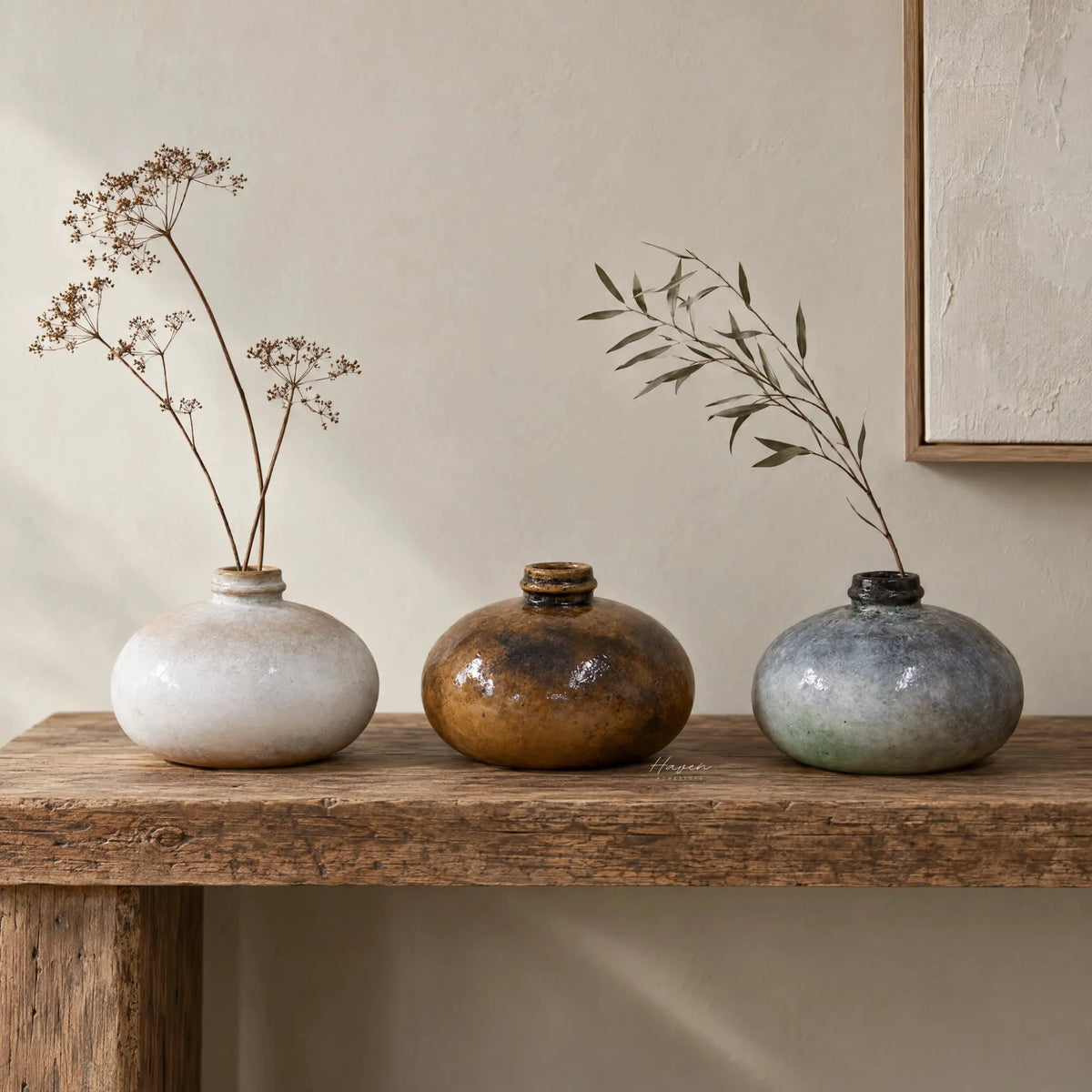 Set of three glossy, low-profile terracotta pots in earthy tones displayed on a wooden console with dried botanical stems.