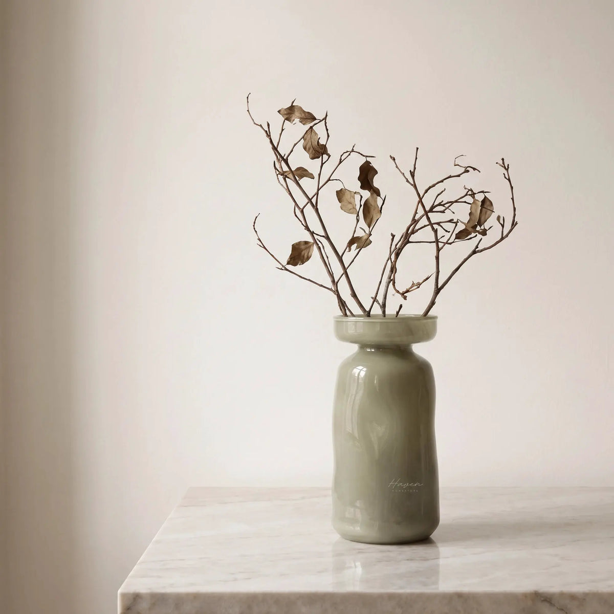 Light green mouthblown glass vase with a flared rim holding dried branches on a marble surface.