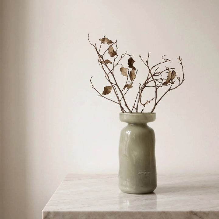 Light green mouthblown glass vase with a flared rim holding dried branches on a marble surface.