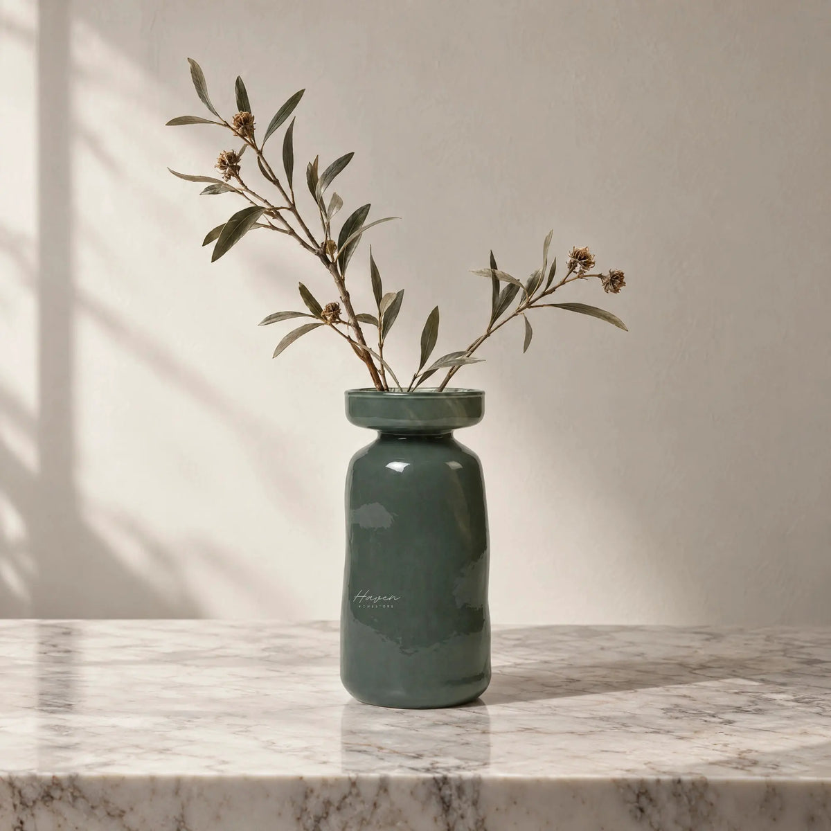 Dark green mouthblown glass vase with a flared rim and rounded body holding dried branches on a marble surface.