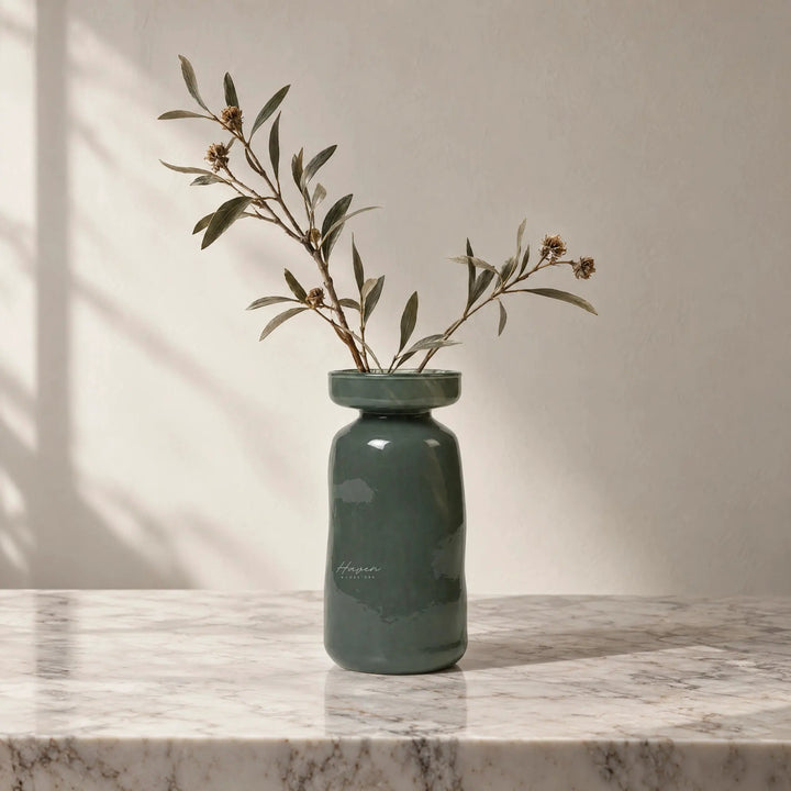 Dark green mouthblown glass vase with a flared rim and rounded body holding dried branches on a marble surface.