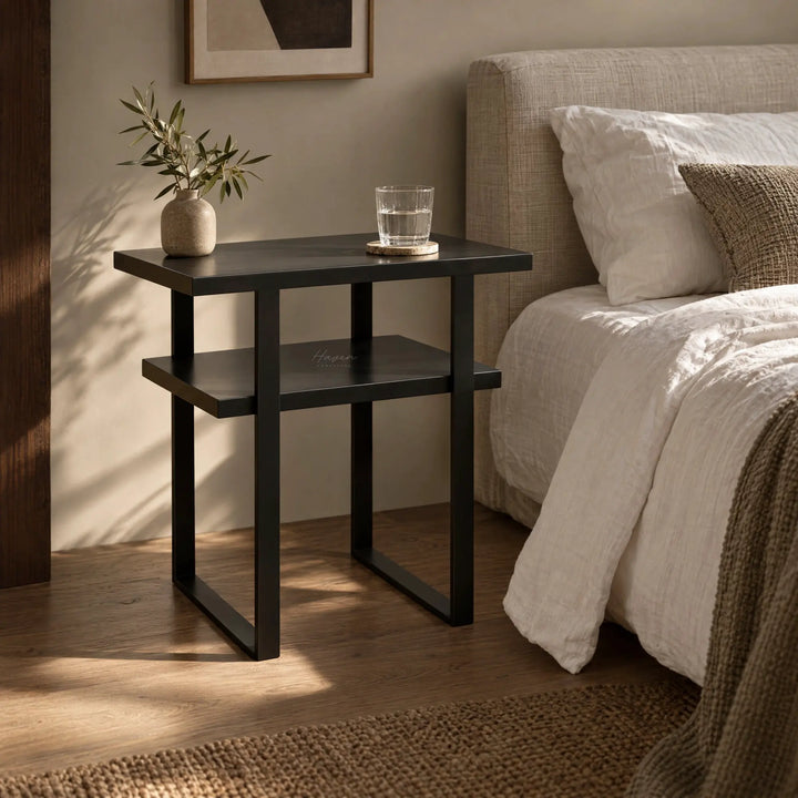 Dark two-tier side table with metal frame beside linen bed, holding vase and glass in sunlit room.