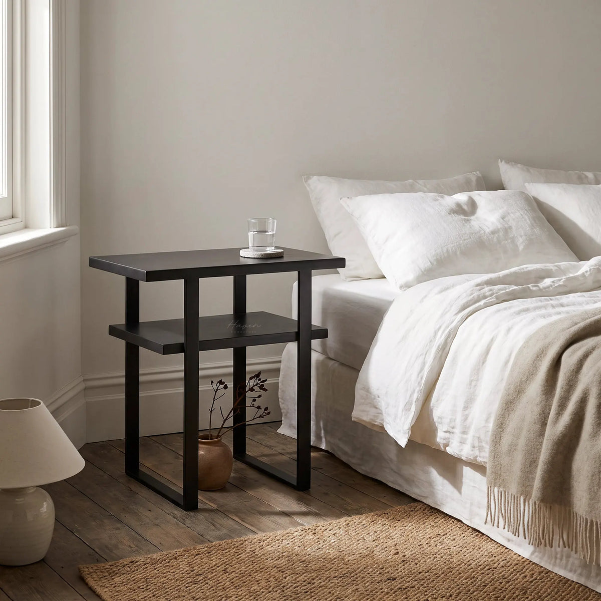Greyed-brown two-tier side table with a simple metal frame stands beside a bed with white linen and a glass of water.
