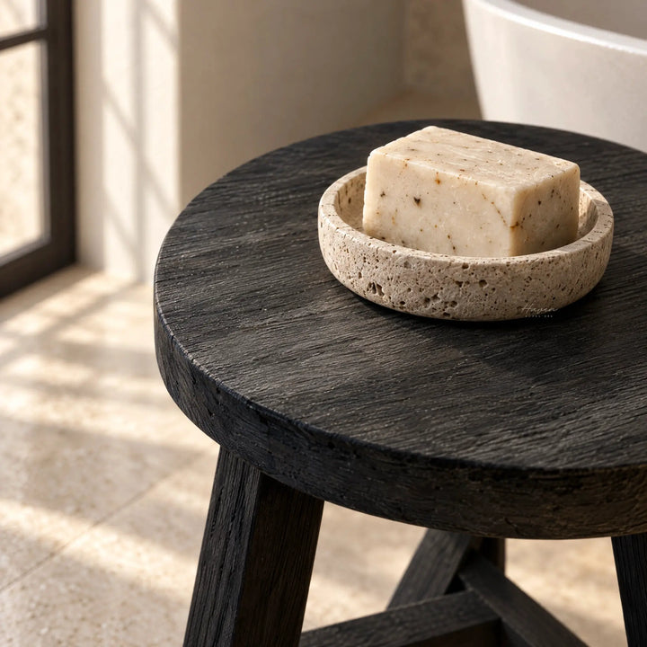 Round black reclaimed elm stool with visible grain holding a travertine soap dish and bar of soap in natural light.