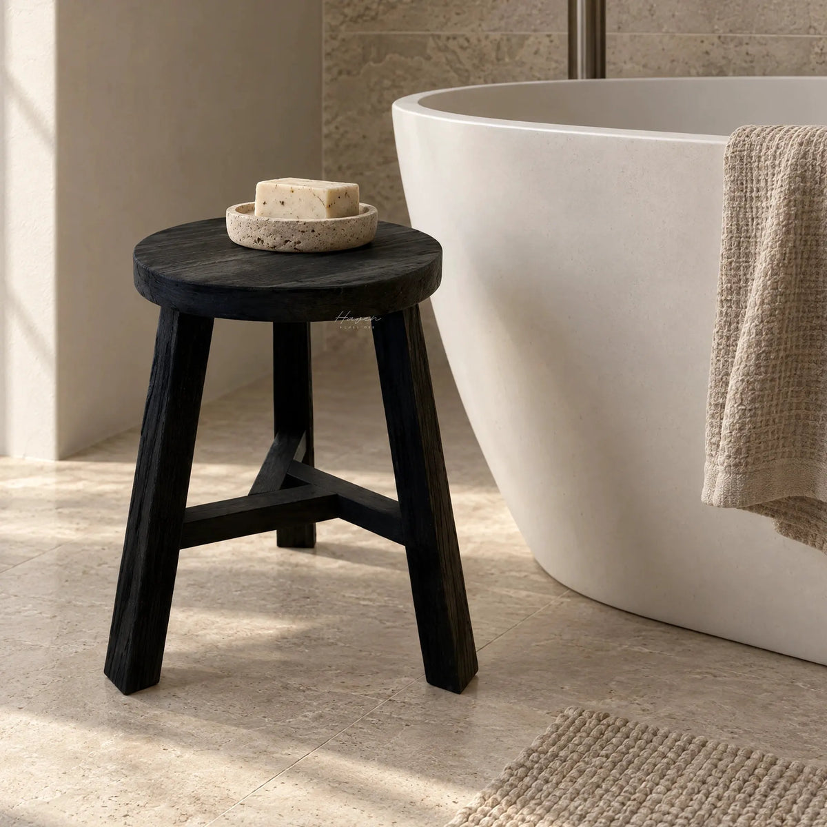 Black reclaimed elm stool with visible grain and knots stands beside a white freestanding tub, holding a stone soap dish.