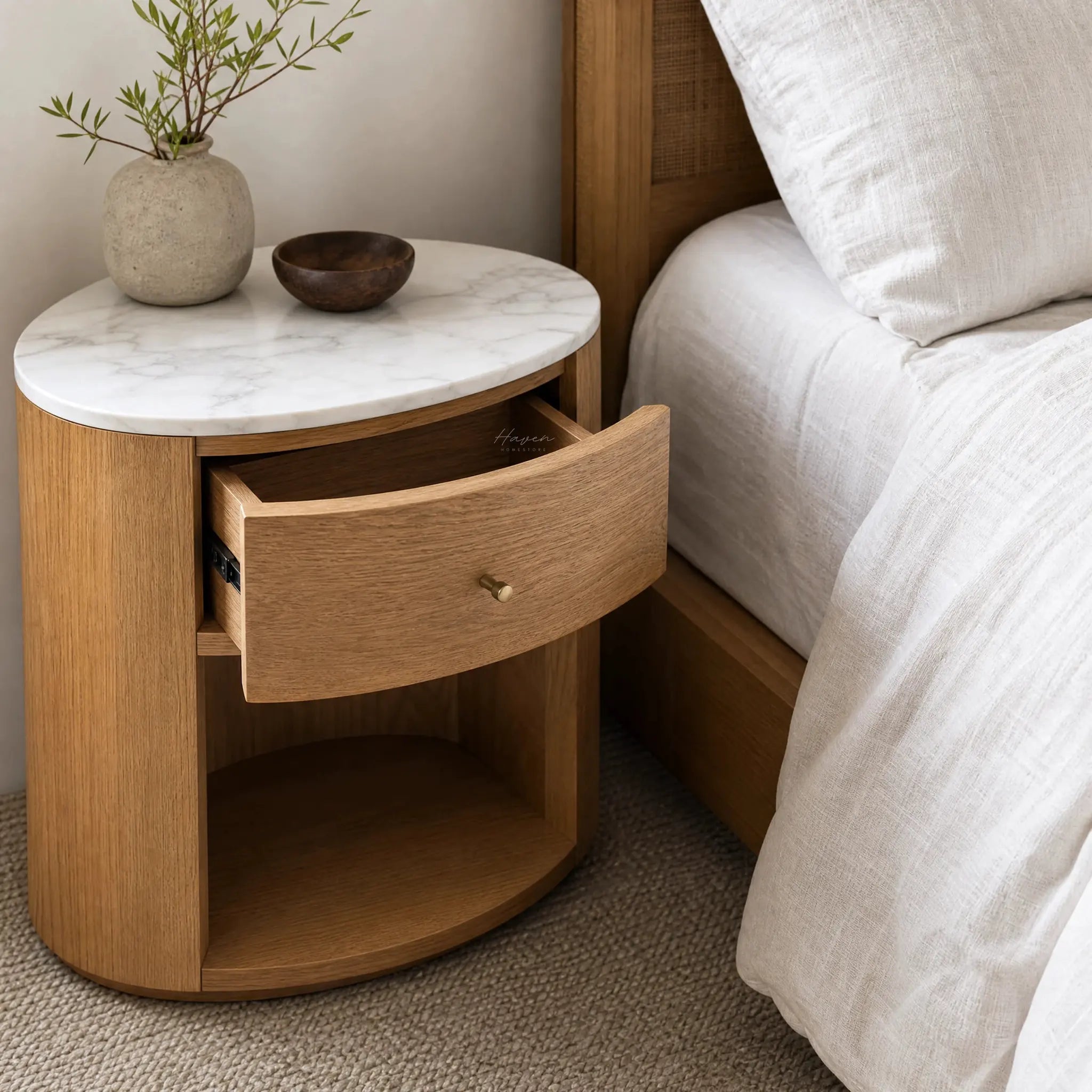 Dana Bedside Table with a marble top and rounded oak frame, open shelf and concealed drawer beside a bed.