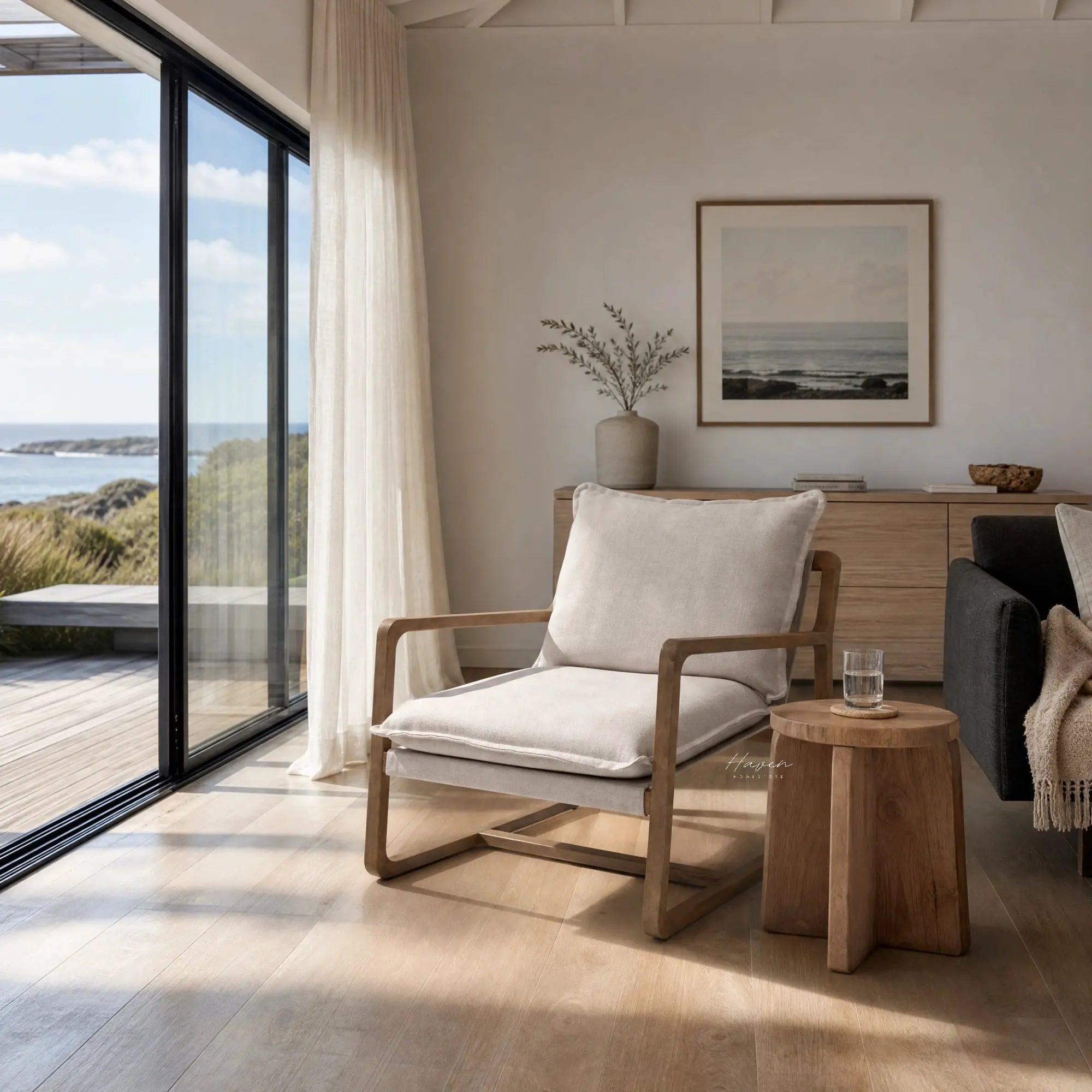 Solid oak lounge chair with soft stone-washed cotton cushions in sand, creating an inviting coastal feel by a large window.