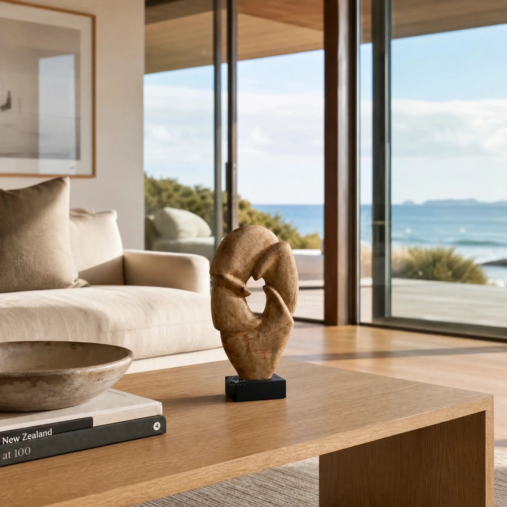 Loop Sculpture in mango wood on a black base, on a wooden console beside a beige sofa, overlooking the coast.