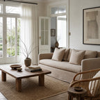 Beige slipcovered sofa with plush cushions in a sunlit living room featuring a rustic wooden coffee table and sheer curtains.